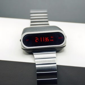 Op de afbeelding: Een zilveren en zwarte digitale polshorloge met een rood digitaal display dat de tijd 2:11:35 PM laat zien. Het horloge heeft een zilveren metalen band en een zwarte wijzerplaat met de woorden "SET", "TIME" en "MODE" erop gedrukt.