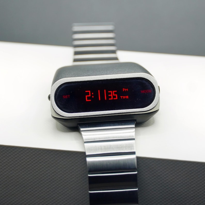 Op de afbeelding: Een zilveren en zwarte digitale polshorloge met een rood digitaal display dat de tijd 2:11:35 PM laat zien. Het horloge heeft een zilveren metalen band en een zwarte wijzerplaat met de woorden "SET", "TIME" en "MODE" erop gedrukt.