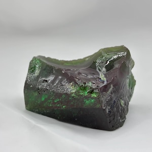 May include: A rough-cut, rectangular gemstone with a dark green and purple color gradient. The stone has a textured surface and appears translucent in areas, with light reflecting off the facets. The overall shape is irregular and natural.