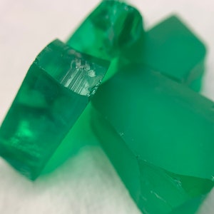 Könnte beinhalten: Nahaufnahme mehrerer smaragdgrüner, unregelmäßig geformter Edelsteine. Die durchscheinenden Steine haben ein poliertes Aussehen mit sichtbaren inneren Strukturen und scharfen, gebrochenen Kanten. Die Edelsteine sind vor einem weißen Hintergrund verstreut.