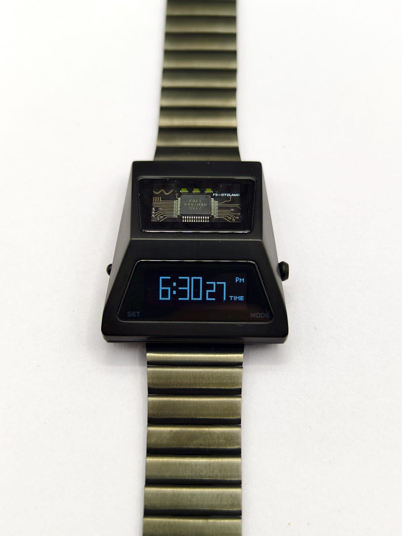 Puede incluir: Un reloj de pulsera digital negro con una correa de metal plateado. La esfera del reloj es de forma triangular con una pantalla digital azul que muestra la hora como 6:30:27 PM. El reloj tiene un bot&oacute;n "SET" en el lado izquierdo y un bot&oacute;n "MODE" en el lado derecho. El reloj tambi&eacute;n tiene una peque&ntilde;a ventana en la parte superior que muestra una placa de circuito.