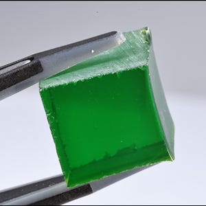 Könnte beinhalten: Ein quadratischer, durchscheinender, smaragdgrüner Edelstein wird von einer grauen Pinzette gehalten. Der Edelstein hat eine leicht raue, unebene Oberfläche und scheint aus einem größeren Stück geschnitten zu sein. Der Hintergrund ist ein weiches, neutrales Grau.