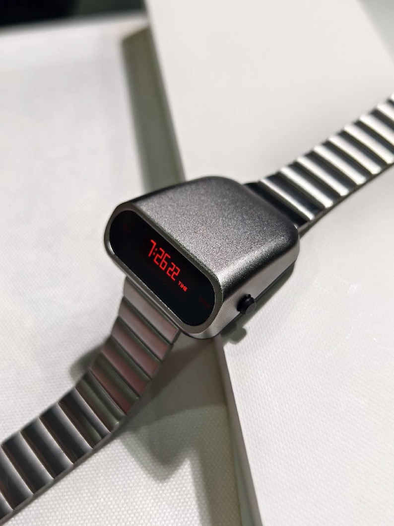 Op de afbeelding: Een zilveren digitale horloge met een rood LED-display dat de tijd 7:26:22 laat zien. Het horloge heeft een metalen band met een geribbeld ontwerp.