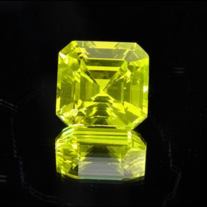 May include: A faceted, emerald-cut, bright yellow gemstone. The gem is set against a black background, with its reflection visible below. The gemstone's facets catch and reflect light, creating a brilliant display. This item is suitable for jewelry making.