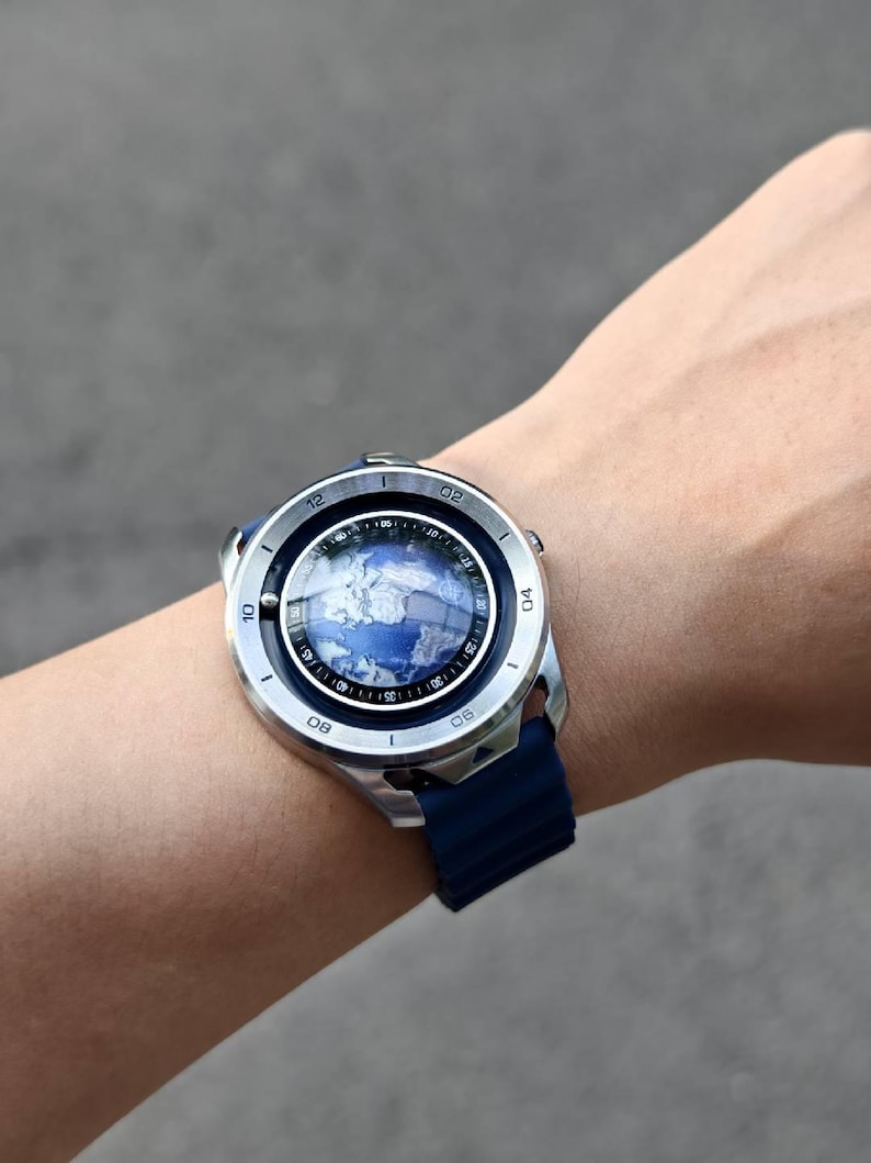 May include: A silver smartwatch with a blue band displays a world map on its screen. The watch has a round face with a blue and white design, and a dark blue silicone strap. The watch is worn on a wrist.