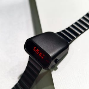 Op de afbeelding: Een zwart digitaal polshorloge met een rood LED-display dat de tijd 18:25:10 uur aangeeft. Het horloge heeft een zwarte metalen band met een geribbeld ontwerp.