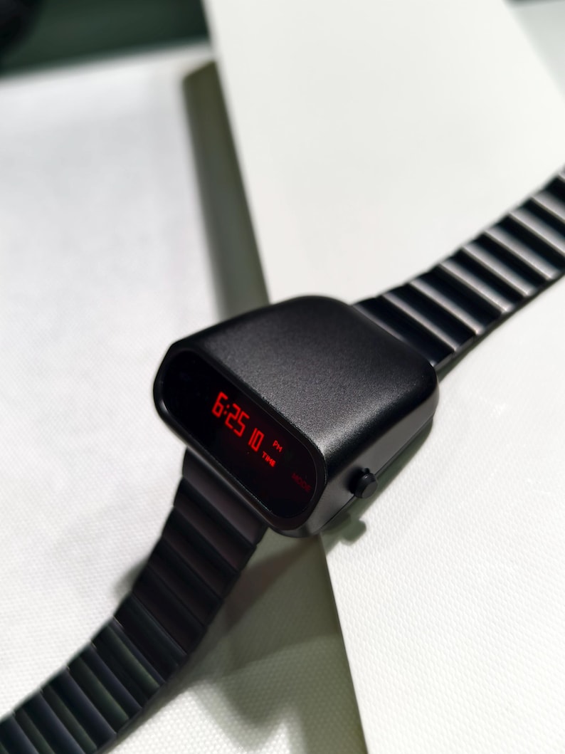 Op de afbeelding: Een zwart digitaal polshorloge met een rood LED-display dat de tijd 18:25:10 uur aangeeft. Het horloge heeft een zwarte metalen band met een geribbeld ontwerp.
