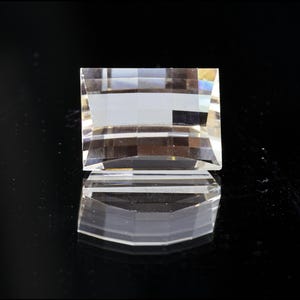 Könnte beinhalten: Ein klares, facettiertes Kristallobjekt mit rechteckiger Form. Der Kristall ist hochreflektierend, mit mehreren Ebenen und Winkeln, die das Licht einfangen. Das Objekt liegt auf einer schwarzen Oberfläche und erzeugt eine Spiegelreflexion.