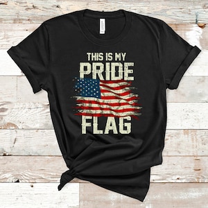 May include: Black t-shirt featuring a distressed American flag design. The shirt has the text "THIS IS MY PRIDE FLAG" in a vintage font. The flag design incorporates red and white stripes with a blue field of stars.