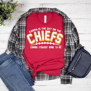 May include: Red t-shirt with a white and yellow graphic that says "Karma is the guy on the Chiefs coming straight home to me". The shirt is laid out on a white wooden surface with a plaid shirt and blue jeans.