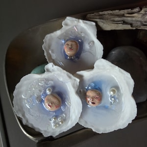 May include: Three white seashell ornaments featuring embedded miniature sculpted faces with painted features. Each shell contains a translucent blue resin pool with small white spherical beads resembling pearls scattered within the glossy surface.