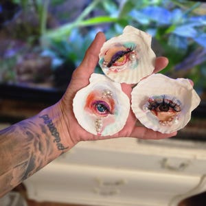 May include: Three white scallop shells each featuring a painted human eye with long black eyelashes. The eyes are surrounded by colorful paint strokes in shades of blue, pink, and orange, accented with small white faux pearls attached to the shells.