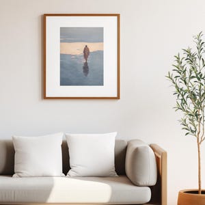 May include: A framed art print depicting a person walking on a beach at sunset. The artwork is in a wooden frame and hangs above a light-colored sofa with white pillows. A potted plant is to the right.