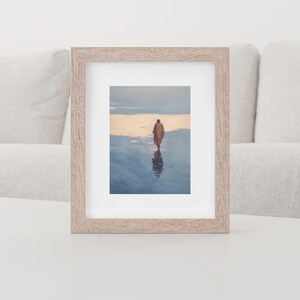 May include: A framed print featuring a person walking on a reflective surface, likely water, with a warm-toned sky in the background. The frame is a light wood color, and the overall aesthetic is serene and contemplative. The image is displayed on a white surface.