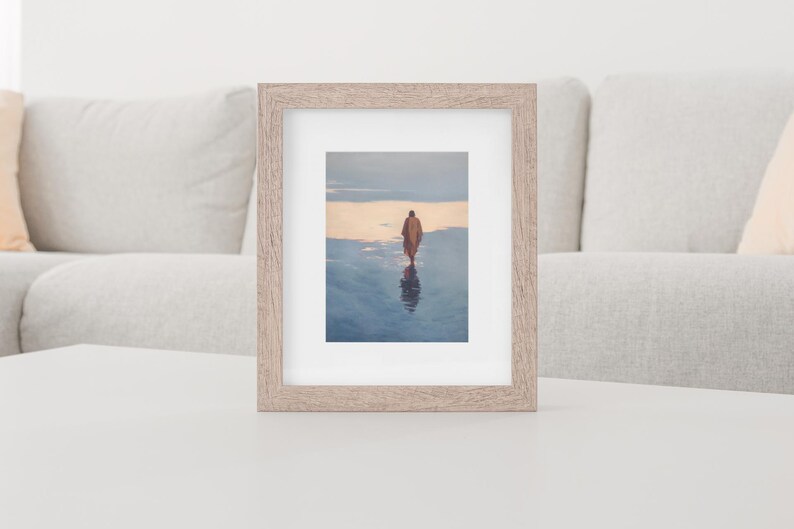 May include: A framed print featuring a person walking on a reflective surface, likely water, with a warm-toned sky in the background. The frame is a light wood color, and the overall aesthetic is serene and contemplative. The image is displayed on a white surface.