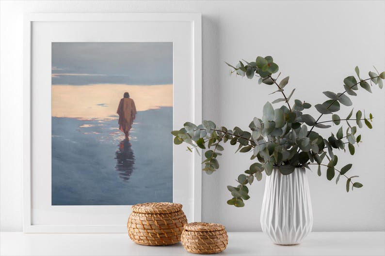 May include: A framed print depicts a figure walking on water, with a warm, golden sky reflected in the water. Two woven baskets and a white vase with greenery complete the scene. The artwork is displayed on a white surface.