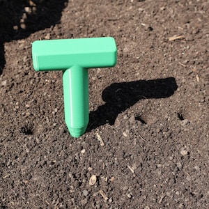 May include: A green plastic hand tool for planting seeds in the garden. The tool has a T-shaped handle and a pointed end.