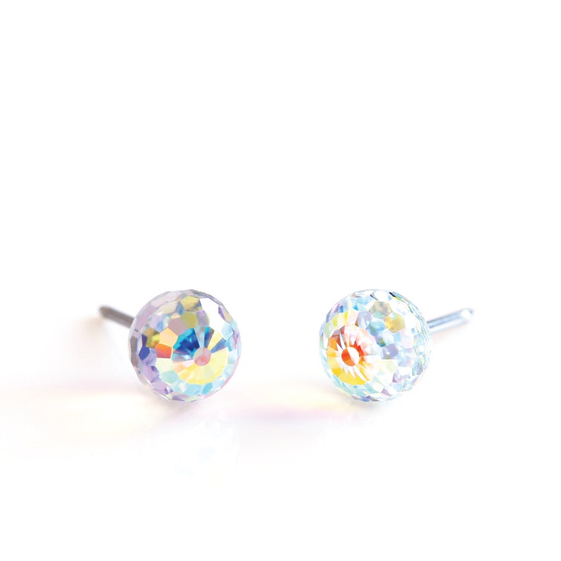 May include: Two iridescent crystal ball stud earrings. Each earring features a faceted, spherical design that reflects a spectrum of colors. The earrings are set on silver posts, creating a minimalist and elegant look.
