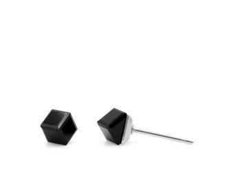 Puede incluir: Un par de pendientes de botón geométricos negros. Cada pendiente presenta una piedra negra facetada en forma de cubo sobre un poste plateado. Los pendientes se muestran sobre un fondo blanco.