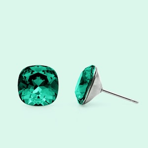 May include: A pair of emerald green cushion-cut stud earrings. The faceted gemstones are set in silver-coloured metal. One earring is viewed from the front, the other from the side, showing the post.