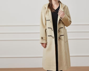 May include: A long, beige hooded coat featuring a front closure with dark brown toggle buttons and loops. The coat has long sleeves with buttoned cuffs, two large rectangular patch pockets on the front, and is worn over a black V-neck top.