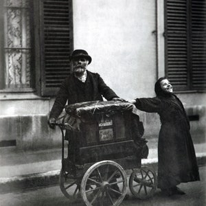Può includere: Una fotografia in bianco e nero di un uomo e una giovane donna che spingono un organo di strada vintage. L'uomo indossa un cappello e un cappotto, e la donna indossa un lungo vestito. L'organo ha un corpo in legno e ruote, ed è decorato con un cartello che dice "Musique".