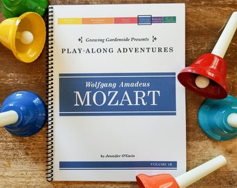 Può includere: Un libro intitolato "Wolfgang Amadeus Mozart" di Jennifer O'Gwin, parte della serie "Play-Along Adventures", con una copertina blu e bianca. Il libro è aperto alla pagina del titolo ed è circondato da campanelle colorate.