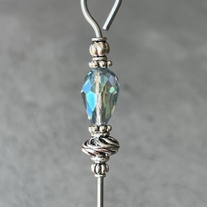 May include: A silver wire with a loop at the top, a silver bead, a light blue iridescent crystal bead, a silver bead with a twisted design, and a silver bead.