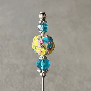 May include: A silver pin with a blue and yellow glass bead with a floral design. The bead is surrounded by two smaller blue glass beads.