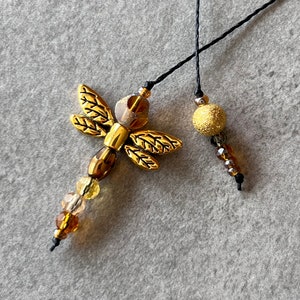 May include: Two gold-toned charms on black cords. One charm is a dragonfly with brown and clear beads. The other charm is a round, sparkly gold bead with brown and clear beads.