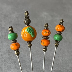 May include: Four decorative metal knitting needles with orange and green glass beads. The beads are shaped like pumpkins with a green leaf design.