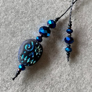 May include: A black and blue beaded necklace with a black stone charm featuring a sun and a swirl design. The necklace is strung on a black cord.