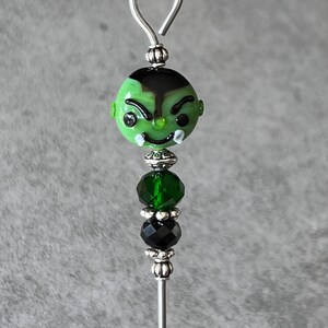 May include: A green glass bead with a cartoon face, black hair, and white teeth. The bead is attached to a silver wire with a loop at the top and three other beads: a silver bead, a green crystal bead, and a black crystal bead.