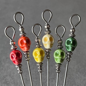 May include: Set of six silver cocktail picks with colorful ceramic skull toppers. The skulls are red, yellow, green, and white.