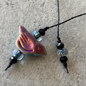 May include: A close-up of a necklace with a iridescent blue bird charm, black beads, and clear iridescent beads. The necklace is strung on black cord.