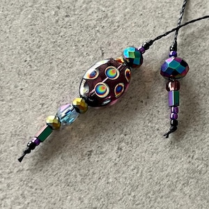 May include: Two beaded pendants with black cords. One pendant features a large, oval, dark purple bead with colorful circles. The other pendant has a faceted, iridescent bead and smaller beads in purple, green, and gold.