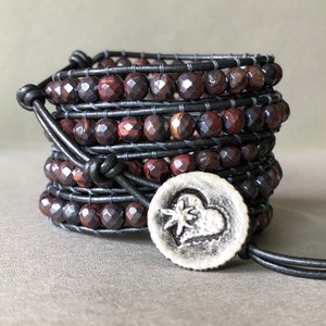 May include: A multi-strand leather wrap bracelet with red and black gemstone beads and a silver charm featuring a heart with a flower inside.