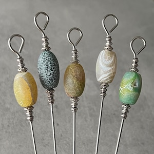 May include: Five decorative cocktail picks with oval-shaped, colorful stone beads. The beads are yellow, green, brown, and white, with silver-colored metal accents and looped tops. These picks are ideal for serving appetizers.
