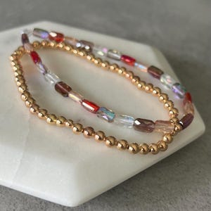 May include: Two bracelets. One is made of small, gold-colored beads. The other bracelet has a mix of colorful, rectangular and iridescent beads. The bracelets are displayed on a white, geometric surface.