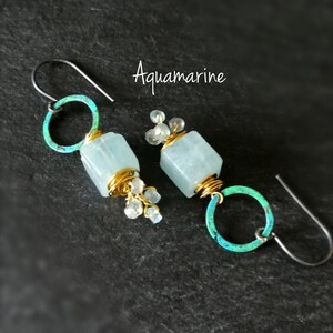 May include: A pair of aquamarine earrings. Each earring features a light blue, cube-shaped stone wrapped in gold wire, with small clear beads and a teal-colored hoop. The word "Aquamarine" is written in cursive.