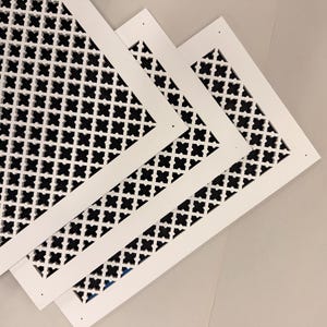 Pode incluir: Três capas de ventilação decorativas brancas com um padrão de trevo preto repetido. As capas são retangulares com uma borda branca e estão dispostas diagonalmente, sobrepondo-se. O fundo é de uma cor cinza claro neutra.