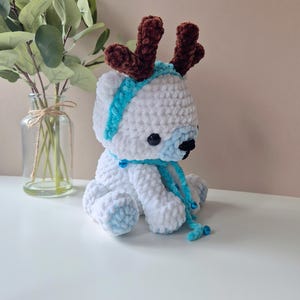 May include: White crocheted plush toy reindeer with brown antlers, a blue scarf, and blue jingle bells.