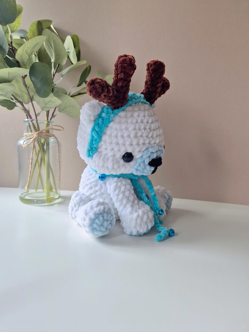 May include: White crocheted plush toy reindeer with brown antlers, a blue scarf, and blue jingle bells.