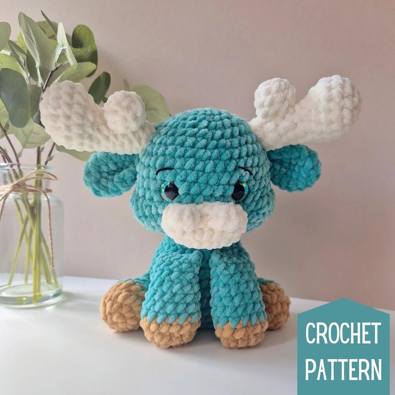 Pu&ograve; includere: Un giocattolo di alce di peluche color turchese lavorato a uncinetto con corna bianche, un naso bianco e zoccoli marroni. Il giocattolo &egrave; seduto su una superficie bianca. CROCHET PATTERN &egrave; scritto in bianco su uno sfondo turchese.