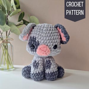 May include: A crocheted gray and pink pig plush toy with black eyes. The pig is sitting with its legs crossed and has a pink nose.
