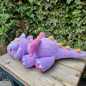 Pu&ograve; includere: Un giocattolo di peluche di drago viola all'uncinetto con accenti rosa e arancioni. Il drago &egrave; sdraiato su una panca di legno.