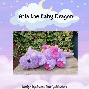 Pu&ograve; includere: Un giocattolo di peluche di drago bambino in crochet viola e rosa con accenti arancioni. Il drago &egrave; sdraiato su una superficie di legno con vegetazione verde sullo sfondo. Il testo "Aria the Baby Dragon" &egrave; sopra l'immagine.