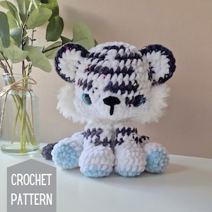 May include: A handmade crochet tiger plushie in white, black, and blue. The tiger has blue eyes, a black nose, and a fluffy white mane. The text "CROCHET PATTERN" is in a gray hexagon shape at the bottom. The plushie is sitting on a white surface.
