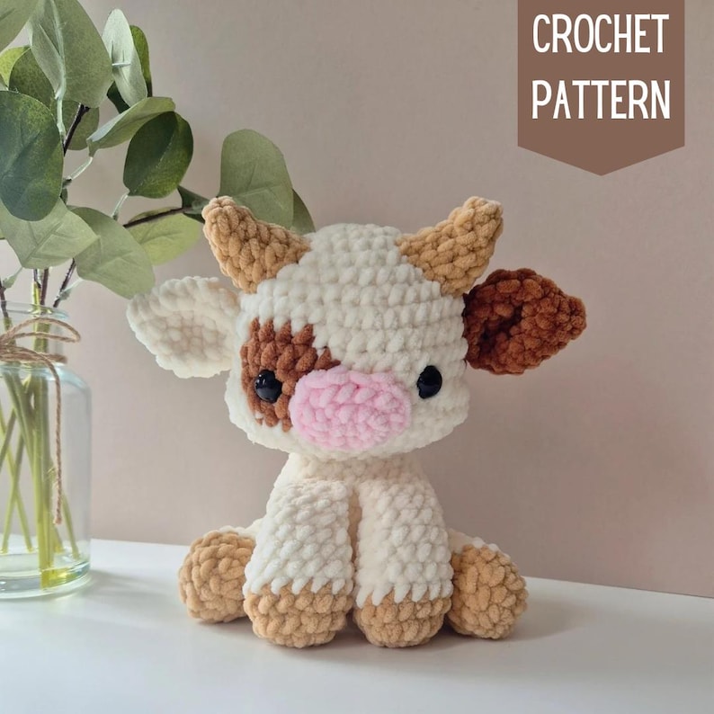May include: Crochet pattern for a white and brown cow amigurumi toy. The cow has a pink nose and black eyes. The pattern is available for download.