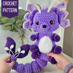 May include: Purple and white crocheted axolotl plush toy with a long, curled tail. The toy has large, black eyes and a white belly.